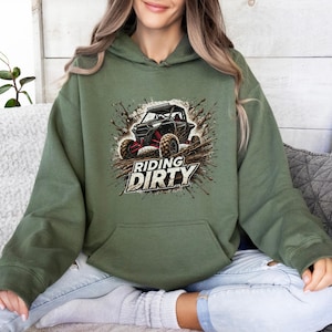 May include: An olive green hoodie featuring a graphic of a black off-road vehicle with red accents, surrounded by a mud splatter design. The words "RIDING DIRTY" are printed below the vehicle in a bold, stylized font.