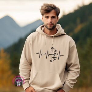 May include: A beige hoodie with a black graphic of a bow and arrow inside a heartbeat. The hoodie has a drawstring hood.