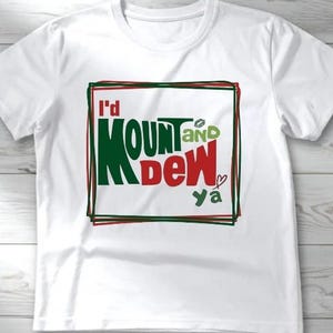 以下が含まれることがあります： 「I'd Mount and Dew Ya」という文字が緑、赤、黒でプリントされた白いTシャツ。テキストは黒と緑の長方形の枠内。小さなハートのグラフィックが「Ya」という単語の隣にあります。