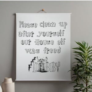 Może przedstawiać: Biały obraz na ścianę z napisem "Please clean up after yourself our House elf was freed" ozdobną czcionką. Obraz zawiera rysunek skrzata domowego Dobby'ego. Na pierwszym planie znajduje się ozdobna roślina i wazony.
