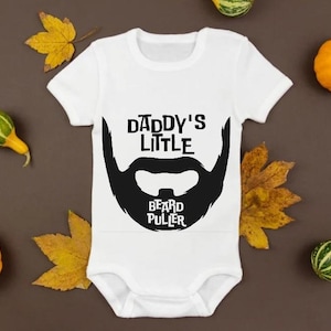 May include: White baby bodysuit with the text "Daddy's Little Beard Puller" in black, featuring a stylized beard graphic. The image is decorated with small pumpkins and fall leaves.