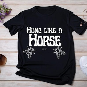 Op de afbeelding: Zwart t-shirt met de witte tekst "Hung Like a Horse" en vliegenillustraties. Het t-shirt wordt gepresenteerd met accessoires zoals een zonnebril en witte schoenen. De tekst is vetgedrukt.