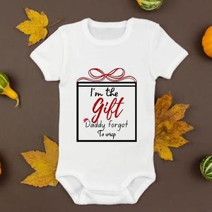 Puede incluir: Body de bebé blanco con el texto "I'm the Gift Daddy forgot to wrap" en rojo y negro. El body está rodeado de pequeñas calabazas y hojas de otoño sobre un fondo marrón.