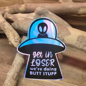 May include: A sticker featuring a black and blue UFO design with an alien silhouette. The text "get in LOSER we're doing BUTT STUFF" is printed in white and pink.
