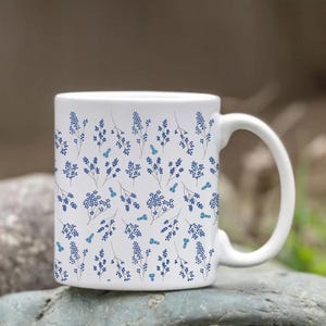 May include: White ceramic mug featuring a repeating pattern of blue floral sprigs and small blue butterfly accents. The mug has a curved handle and is set against a blurred natural background, suggesting outdoor use.