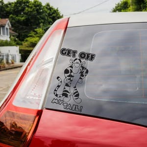 Può includere: Un adesivo in bianco e nero con Tigro di Winnie the Pooh con la scritta "GET OFF MY TAIL!". L'adesivo è sul lunotto posteriore di un'auto rossa.