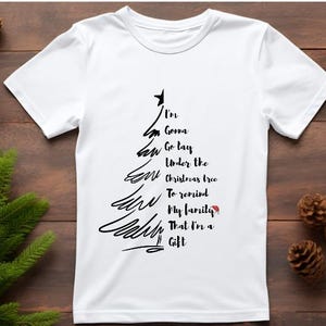 May include: White t-shirt with a black Christmas tree design and the text "I'm Gonna Go Lay Under the Christmas tree To remind My family That I'm a Gift". The shirt is on a wooden surface with pine cones and greenery.