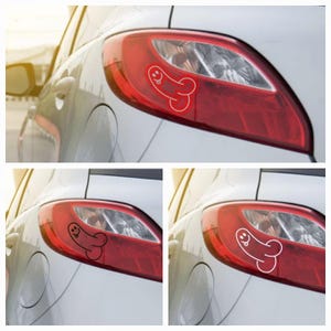 Puede incluir: Un coche plateado con un piloto trasero rojo. Una pegatina blanca de un pene de dibujos animados con una cara sonriente está aplicada al piloto trasero.