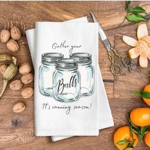 May include: A white kitchen towel with a watercolor illustration of three mason jars with the text "Gather your Balls" and "It's canning season!". The towel is on a wooden surface with walnuts, tangerines, and pine branches.