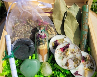 May include: A wooden box filled with various items, including a small glass bottle with herbs and crystals, two green stones, a black charcoal disc, a white candle, a white paper scroll, and bay leaves. The box is lined with green shredded paper.