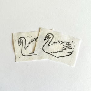 May include: Two rectangular off-white fabric patches featuring a black ink block print of a swan. Each patch displays a stylized, hand-drawn outline of a swan with a long neck and feathered wings, rendered in a simple, bold, and graphic line style.