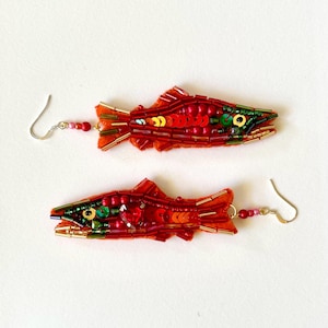 Pode incluir: Um par de brincos artesanais em forma de peixe. Os brincos são predominantemente vermelhos com detalhes em verde, dourado e laranja. São adornados com contas e lantejoulas e possuem ganchos prateados.
