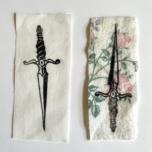 Op de afbeelding: Twee rechthoekige stoffen patches met dolkillustraties. De linker patch is effen gebroken wit met een zwarte inkt dolk. De rechter patch heeft een bloemenpatroon met roze rozen en groene bladeren, bedrukt met een zwarte inkt dolk.