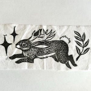 May include: A rectangular piece of off-white fabric featuring a black ink block print of a jackalope in motion. The design includes two four-pointed stars on the left side and a leafy branch on the right side, all rendered in a bold, graphic style.