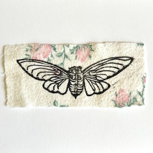 May include: A rectangular fabric patch featuring a black ink illustration of a cicada centred on a cream-coloured background with a repeating pattern of small pink rosebuds and green leaves with frayed, uneven edges on all sides.