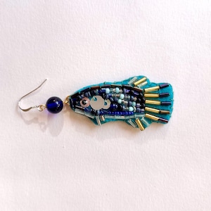 Op de afbeelding: Een handgemaakte, turquoise visvormige oorbel met ingewikkeld kralenwerk. De oorbel heeft een zilveren vishaak, een donkerblauwe kraal en een lichaam versierd met blauwe, zilveren en gouden kralen. De vis heeft een paillettenoog en een vilten achterkant.
