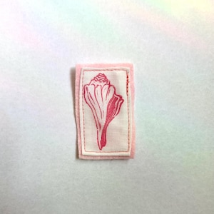 May include: A pink fabric patch with a seashell design. The seashell is printed in a darker shade of pink on a white background. The patch is rectangular with a pink border and white stitching.