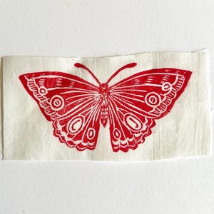 May include: A rectangular piece of off-white fabric featuring a centered, bold red ink print of a butterfly. The butterfly design displays intricate line work, symmetrical wing patterns, and circular eye-like markings on each of the four wings.