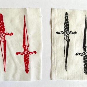 Op de afbeelding: Twee rechthoekige stoffen patches met blokdrukontwerpen van dolken. De linker patch toont twee rode dolken, terwijl de rechter patch twee zwarte dolken bevat. Elke dolk heeft een gedetailleerd, gedraaid handvat en een scherp, puntig blad.