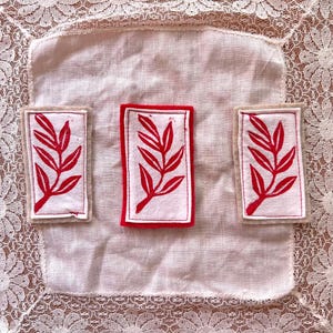 May include: Three rectangular fabric patches with a red leaf design on a white background. The patches are framed with either a red or beige border. The patches are arranged on a white cloth with a lace border.