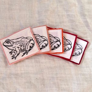 May include: A set of five square fabric coasters with a frog illustration. Each coaster has a white center with a black frog design and a colored border in shades of pink, orange, and red. The coasters are arranged on a light-colored fabric surface.