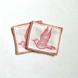 May include: Two square fabric patches with a pink bird design. The bird is printed in pink ink on a white background. The patches are bordered with a pink felt material.