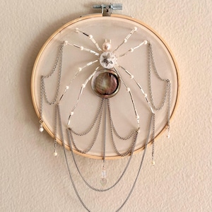Op de afbeelding: Een decoratieve spin geborduurd op een houten borduurring. De poten zijn gemaakt van kralen en zilveren kettingen. Het lichaam is versierd met een parelmoer schelp en hangende kettingen. Een hartvormige bedel hangt eraan.