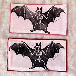Pode incluir: Duas aplicações retangulares com um desenho de esqueleto de morcego preto. Os morcegos têm as asas abertas e são impressos em tecido branco. Uma aplicação tem uma borda rosa, a outra uma borda bordô.