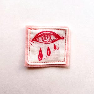 Pode incluir: Um patch quadrado com um design de olho vermelho e lágrimas sobre um fundo branco, colocado sobre um quadrado de feltro rosa. O olho é detalhado com uma íris e cílios estilizados. O patch é provavelmente para roupas ou acessórios.