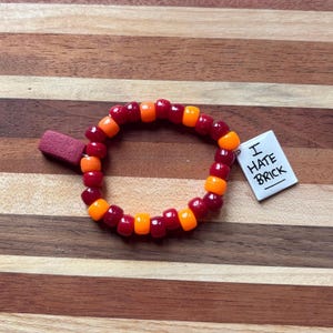 Pode incluir: Uma pulseira circular feita de contas de plástico alternadas em vermelho escuro e laranja brilhante. A pulseira apresenta um pequeno pingente retangular vermelho e um pingente quadrado branco com o texto preto I HATE BRICK impresso.