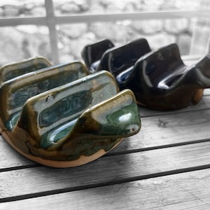 May include: Two ceramic taco holders with a glossy finish. One is green and brown, the other is dark blue. Each holder has three curved slots to hold tacos upright. They sit on a light-colored wooden surface.