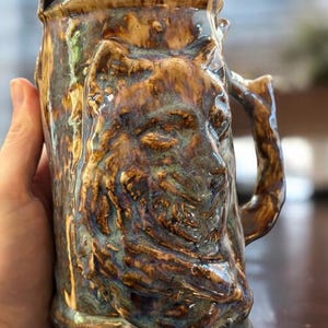 Könnte beinhalten: Eine Keramiktasse mit einem Wolfskopf, der auf der Vorderseite modelliert ist. Die Tasse ist in braunen, grünen und blauen Tönen glasiert.