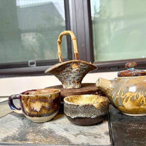 May include: A ceramic tea set with a teapot, cup, bowl, and a basket-shaped container. The teapot and cup have brown, blue, and yellow glaze. The basket has a bamboo handle. The bowl is brown and white.