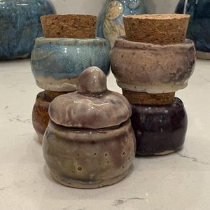 May include: A selection of small, handmade ceramic jars with cork stoppers. The jars are in various colours, including blue, brown, and purple, with a glossy finish. One jar has a lid with a rounded knob. These decorative jars are ideal for storing small items.