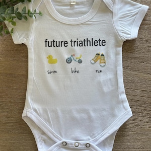 Könnte beinhalten: Weißer Baby-Body mit dem Text "future triathlete" und Illustrationen einer gelben Ente, eines Fahrrads und Laufschuhen. Der Body hat die Größe 0-3M.