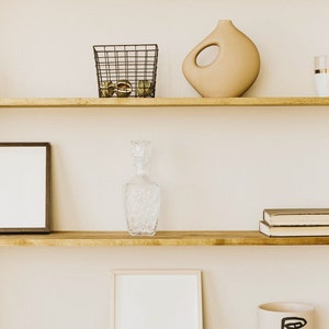 May include: Wooden shelves with various decorative items. Includes a framed picture, a glass decanter, a beige vase with a hole, a candle, a mug, a wire basket, and books. The shelves are light brown and the background is a neutral color.