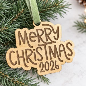 Peut inclure: Une décoration en bois clair aux bords festonnés portant l'inscription gravée Merry Christmas 2026. L'ornement est suspendu par un ruban de velours vert sauge, mettant en valeur un grain de bois naturel et des lettres gravées sombres.