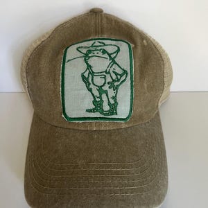 Pode incluir: Boné de beisebol bege com costas em rede e aba curva. A frente apresenta um patch retangular com contorno verde e um sapo de desenho animado usando macacão e chapéu. O boné é de cor neutra.