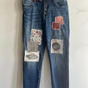 May include: Blue denim jeans featuring a button-fly closure and various fabric patches sewn onto the front. The patches include a floral print, a moth illustration, a spiral design, and solid-colored squares in shades of pink and brown.