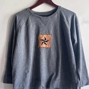 May include: A heather gray long-sleeve sweatshirt featuring a square tan fabric patch stitched onto the center chest. The patch displays a black five-pointed star graphic with visible dark thread stitching around the perimeter of the square.