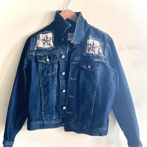 May include: A dark blue denim jacket featuring a pointed collar, long sleeves, and a button-up front. Two chest pockets with buttoned flaps are present. Each shoulder area is embellished with a square patch featuring a black star design.