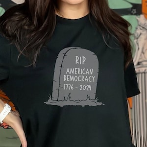 May include: A black t-shirt with a gray graphic of a tombstone that reads "RIP American Democracy 1776-2024".