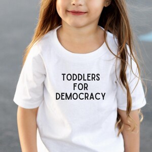 May include: A young child wearing a white t-shirt with the text "TODDLERS FOR DEMOCRACY" in black. The child is smiling and wearing black shorts. The background is a gray surface.