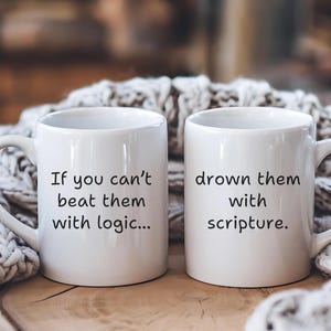 May include: Two white ceramic mugs with black text. One mug reads "If you can't beat them with logic..." and the other reads "drown them with scripture." The mugs are on a wooden surface with a knitted blanket in the background.
