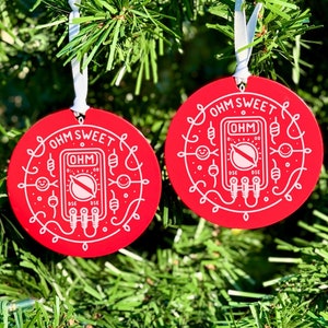 May include: Two red, round Christmas ornaments with white text and illustrations. Each ornament says "OHM SWEET" and features a multimeter design. They hang from white ribbons against a backdrop of green Christmas tree branches.