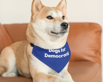 May include: A Shiba Inu dog wearing a blue bandana with the white text "Dogs for Democracy." The dog is light brown and white, and is sitting on a brown leather couch. The dog is looking to the right.