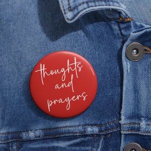 May include: A red button with white text that reads "thoughts and prayers". The button is pinned to a blue denim jacket.