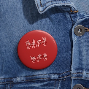 May include: A round, red button with white hand gestures spelling out the word "Deaf". The button is set against a blue denim jacket.