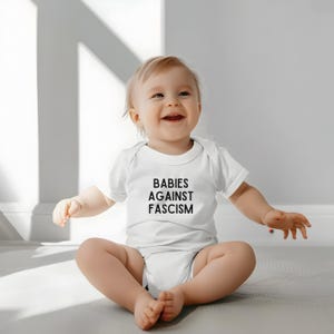 May include: A smiling baby wearing a white onesie with the text "BABIES AGAINST FASCISM" in black. The baby is sitting on a light-colored surface, with arms and legs outstretched.