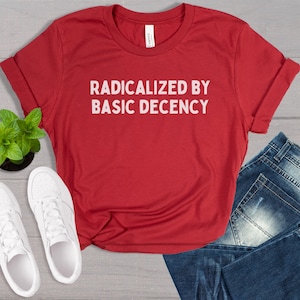 May include: Red t-shirt with white text that reads "Radicalized by Basic Decency". The shirt is laid flat on a wooden surface with a pair of white sneakers and blue jeans.